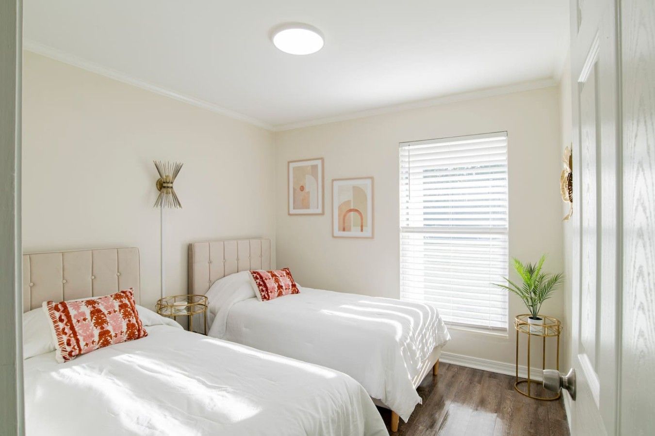 A bedroom with two beds, a bedside table, a floor lamp, a window with blinds, and a small plant.