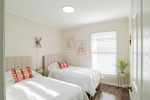 A bedroom with two beds, a bedside table, a floor lamp, a window with blinds, and a small plant. - South Alabama Home near Colleges (Mobile)