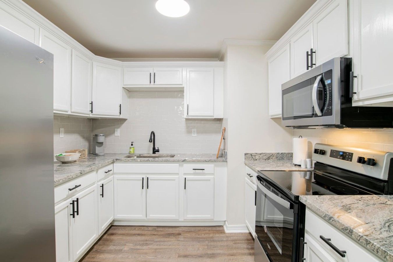 Una cocina moderna con armarios blancos, encimera de granito, refrigerador de acero inoxidable, microondas y horno empotrado.