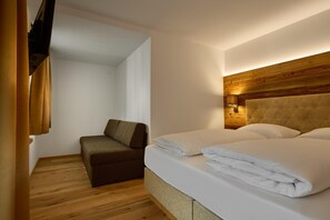 A hotel room with a wooden headboard, a bed with white linens, a brown sofa, and a wooden floor. - Modern Suite With Panoramic Views (Gaschurn)