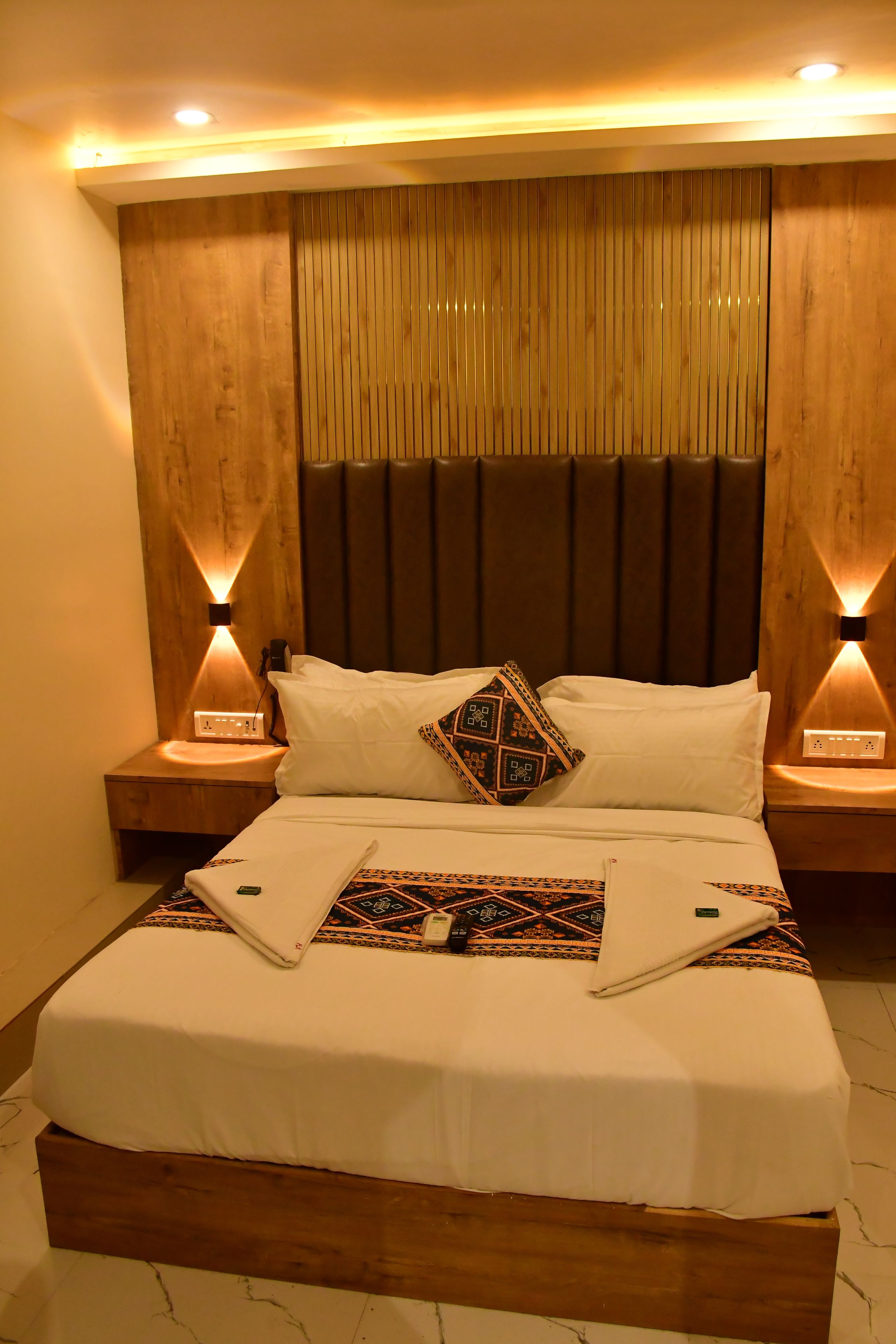 Habitación de hotel con cabecera de madera, dos lámparas de noche, cama con ropa blanca y un cojín decorativo.