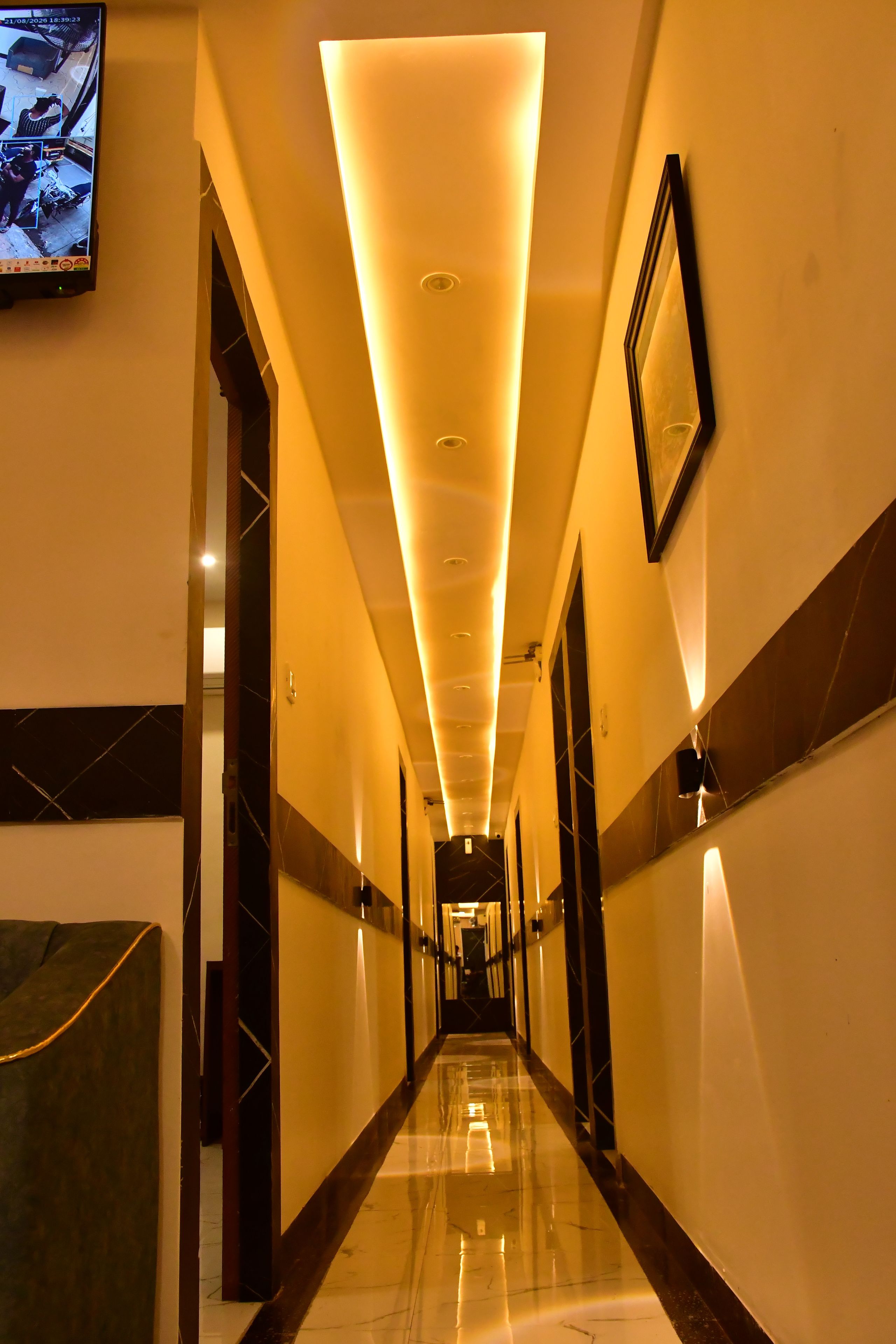 A hotel corridor with a polished floor, recessed lighting, and a television screen displaying a security feed.