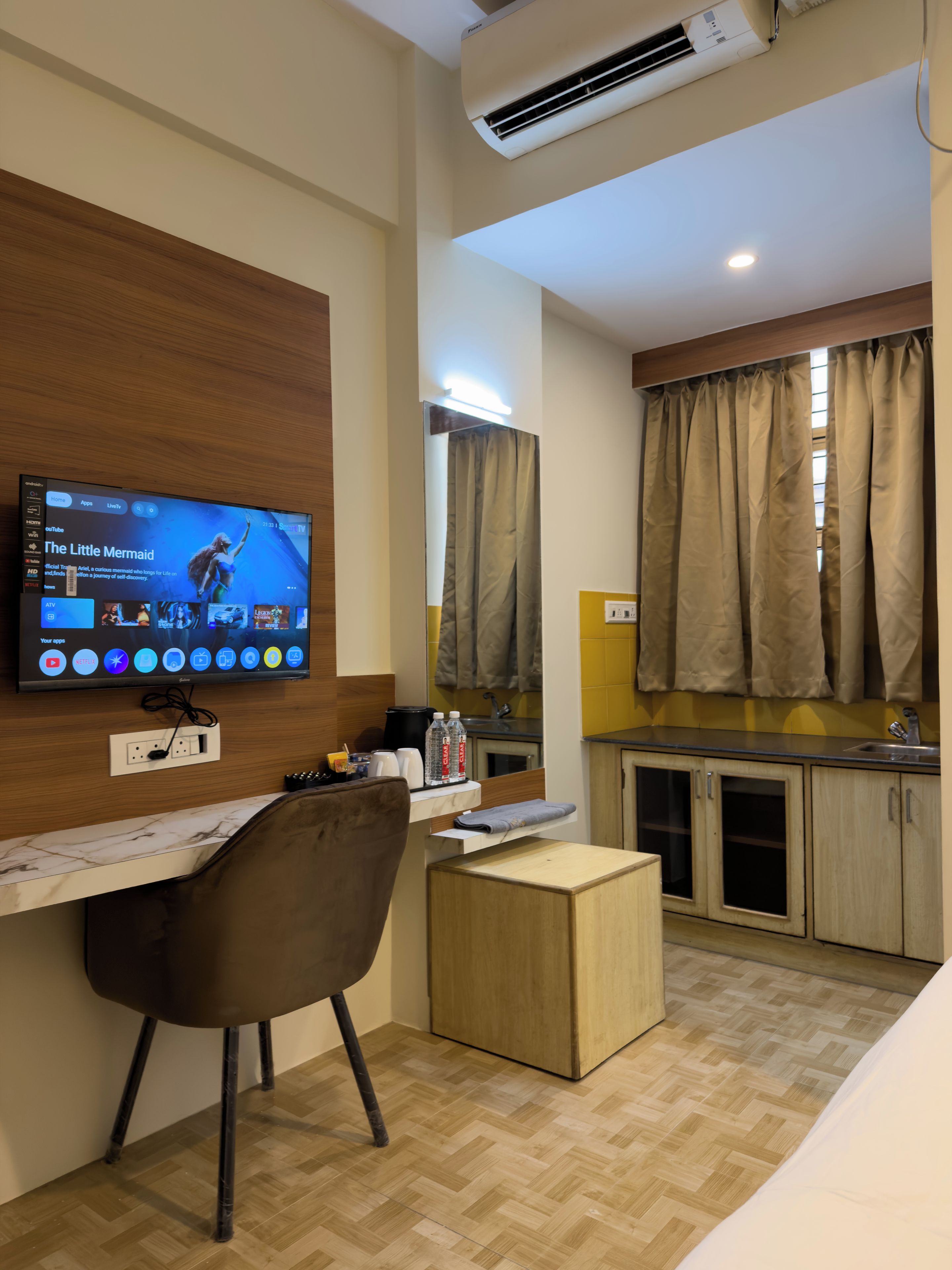 A hotel room with a TV on the wall, a desk, a chair, a small wooden table, a kitchen area with cabinets, and a window with curtains.