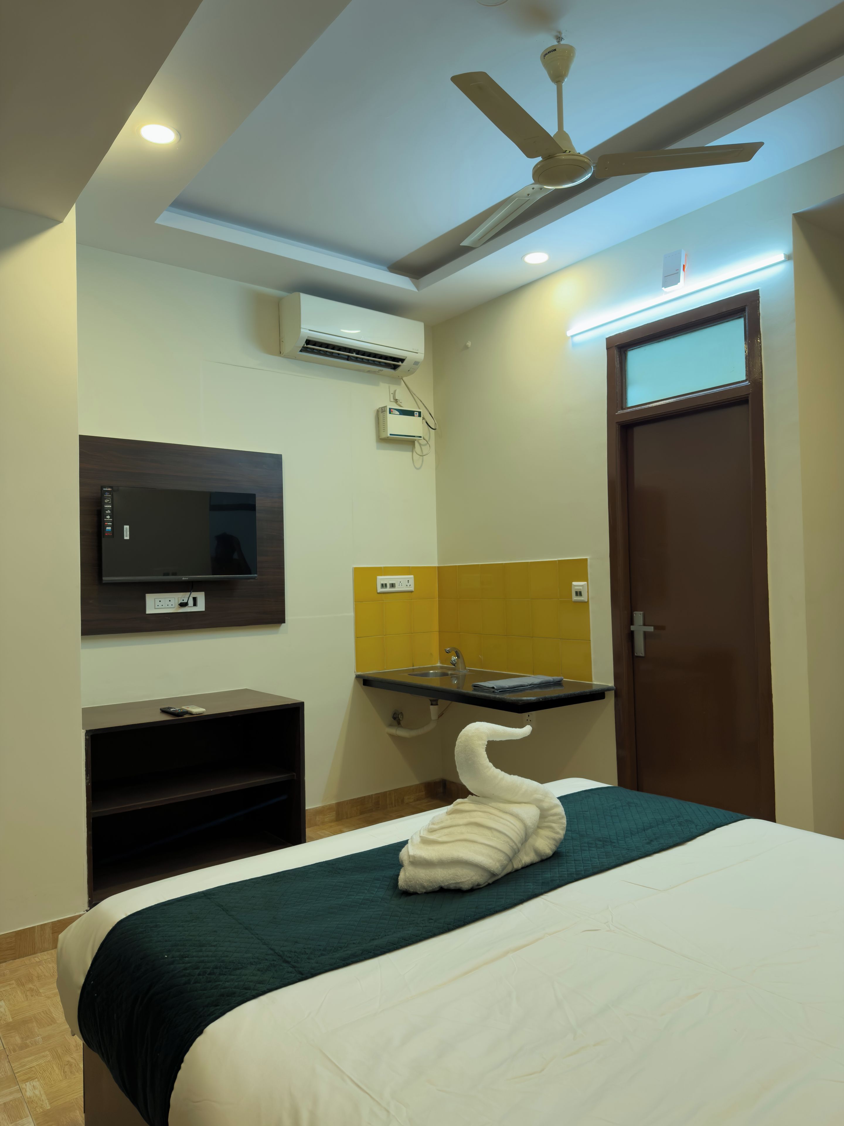 Hotelkamer met een bed, een flatscreen televisie, een airconditioner, een plafondventilator, een badkamer met een wastafel en een deur.