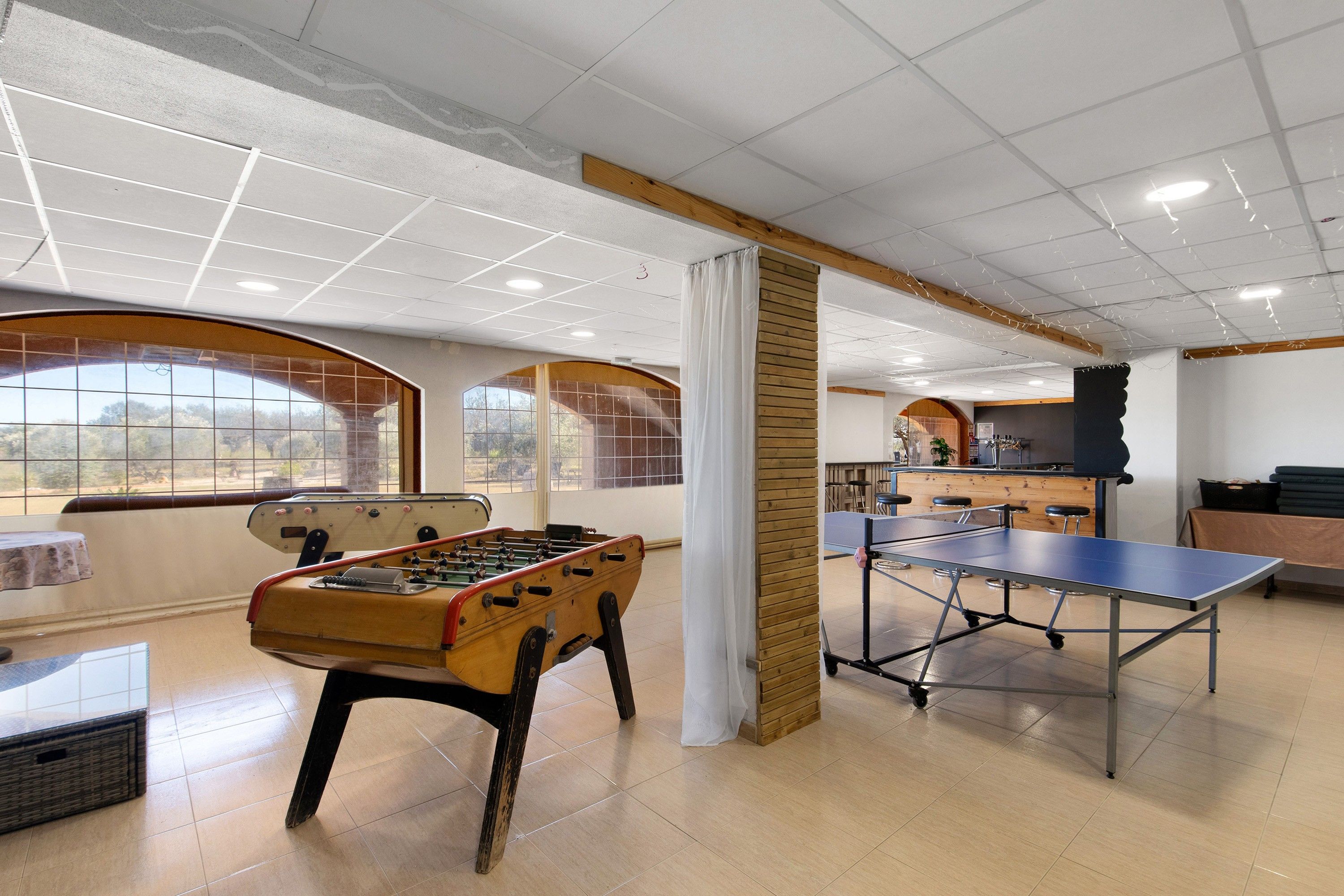 A room with a foosball table, ping pong table, and a bar area with stools.