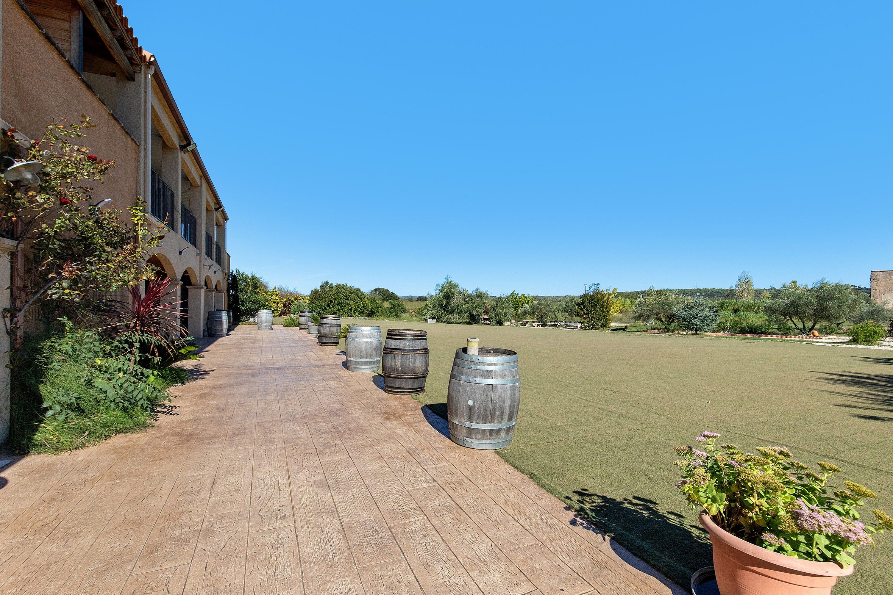 A pathway lined with large barrels, a building with arched windows, and a grassy field.
