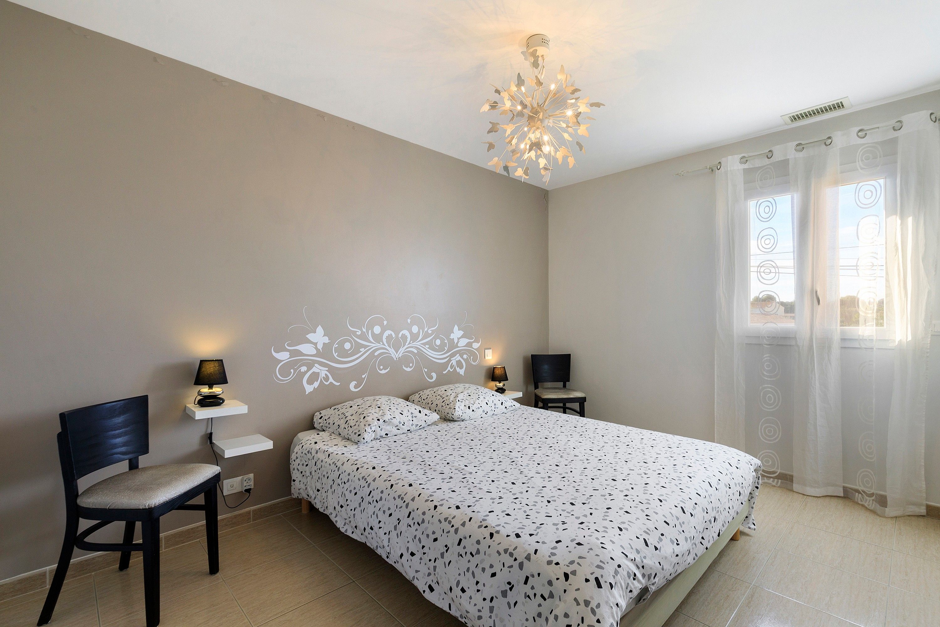 A bedroom with a bed, two bedside tables with lamps, a chair, a decorative ceiling light, and a window with curtains.