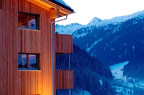 A wooden building with multiple windows, set against a backdrop of snow-covered mountains and a forested valley. - Chalet With Fireplace and Balcony (Gaschurn)