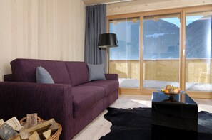 A room with a purple sofa, a black lamp, a wooden table with fruit, and a view of mountains through a large window. - Chalet With Fireplace and Balcony (Gaschurn)