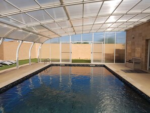 Una piscina interior con techo de vidrio, una banca y vista a un área ajardinada.