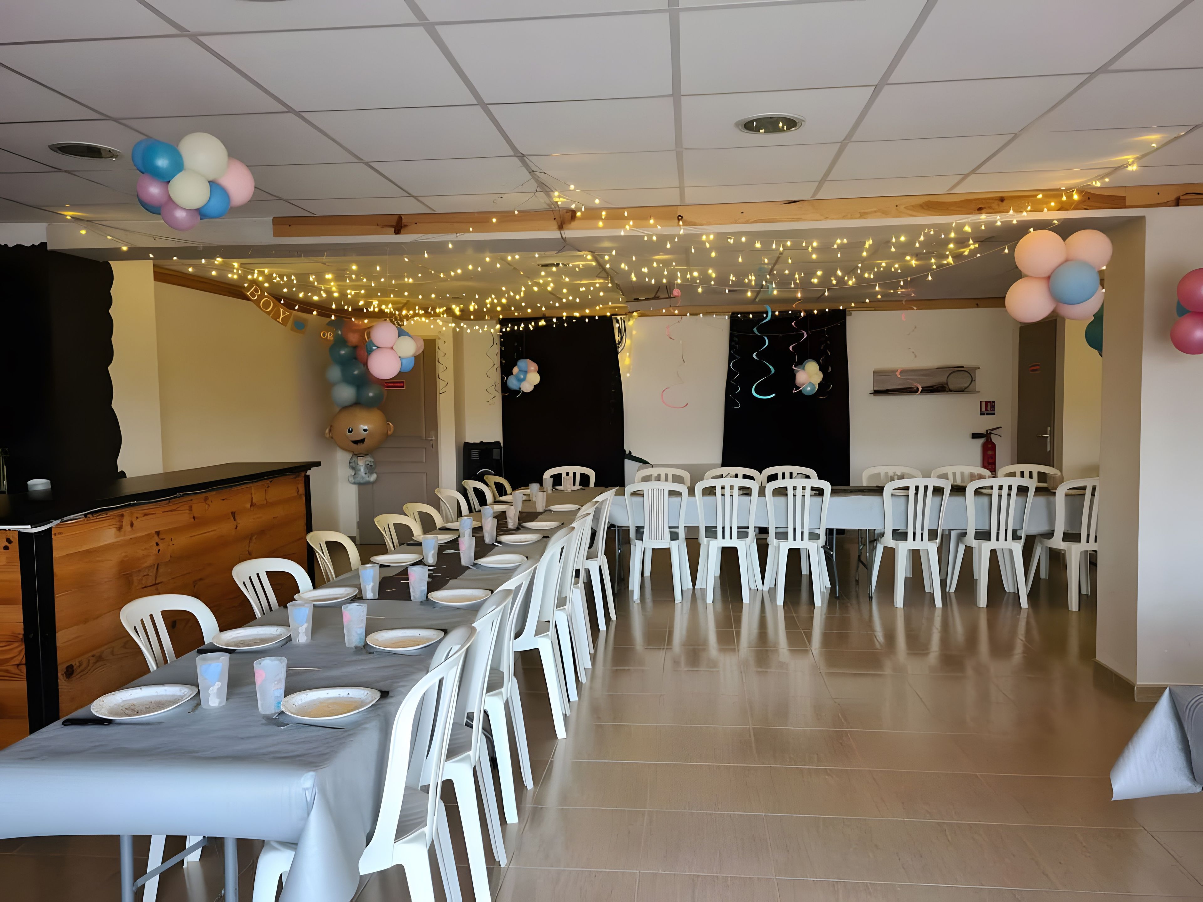 A long dining table with white chairs, surrounded by walls with ceiling lights and balloons.