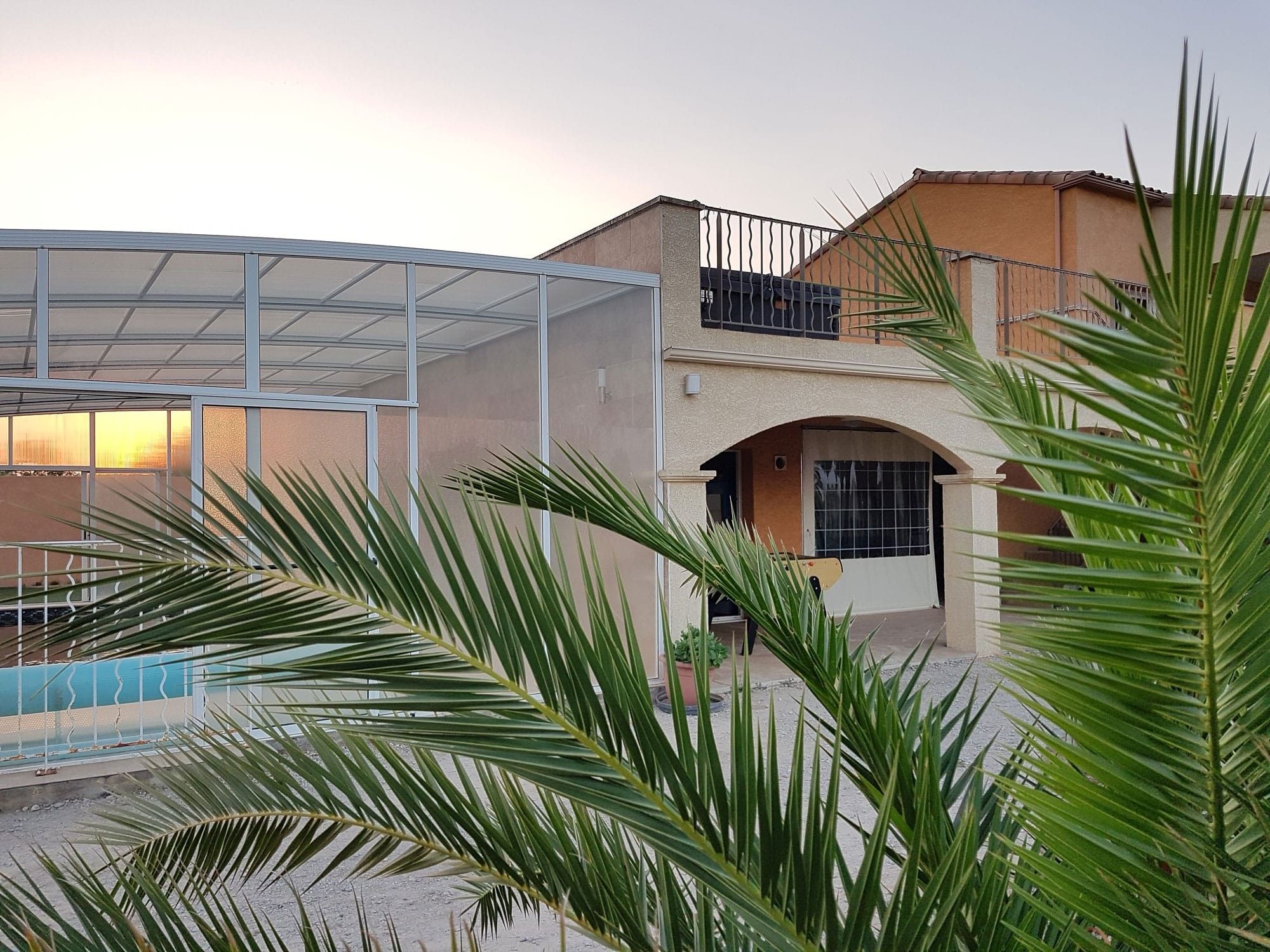A residential building with a covered swimming pool, a balcony with a metal railing, and palm trees in the foreground.