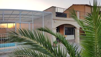 A residential building with a covered swimming pool, a balcony with a metal railing, and palm trees in the foreground.
