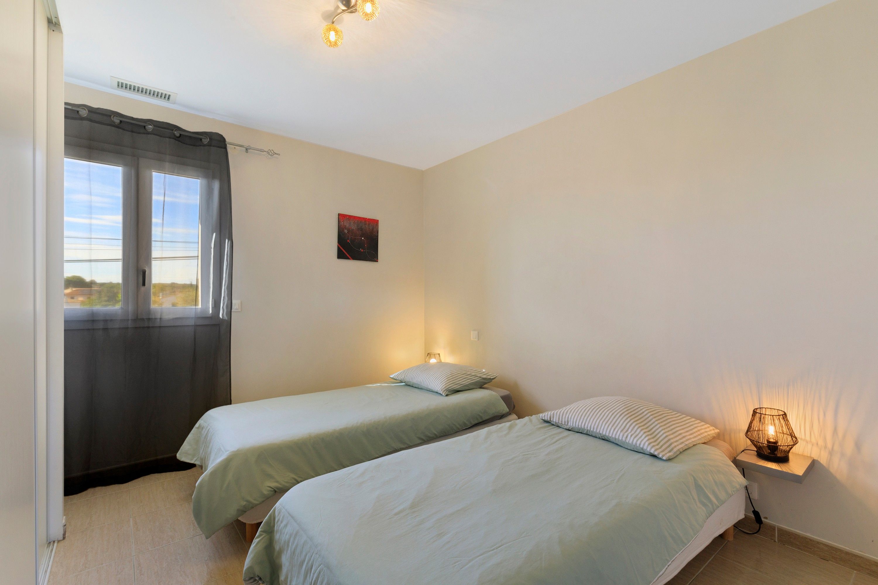 A bedroom with two beds, a window with curtains, a wall-mounted artwork, and a bedside lamp.