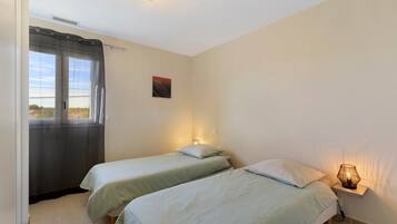 A bedroom with two beds, a window with curtains, a wall-mounted artwork, and a bedside lamp.