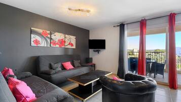 A modern living room with a grey sofa, a black ottoman, a coffee table, and a flat-screen TV mounted on the wall.