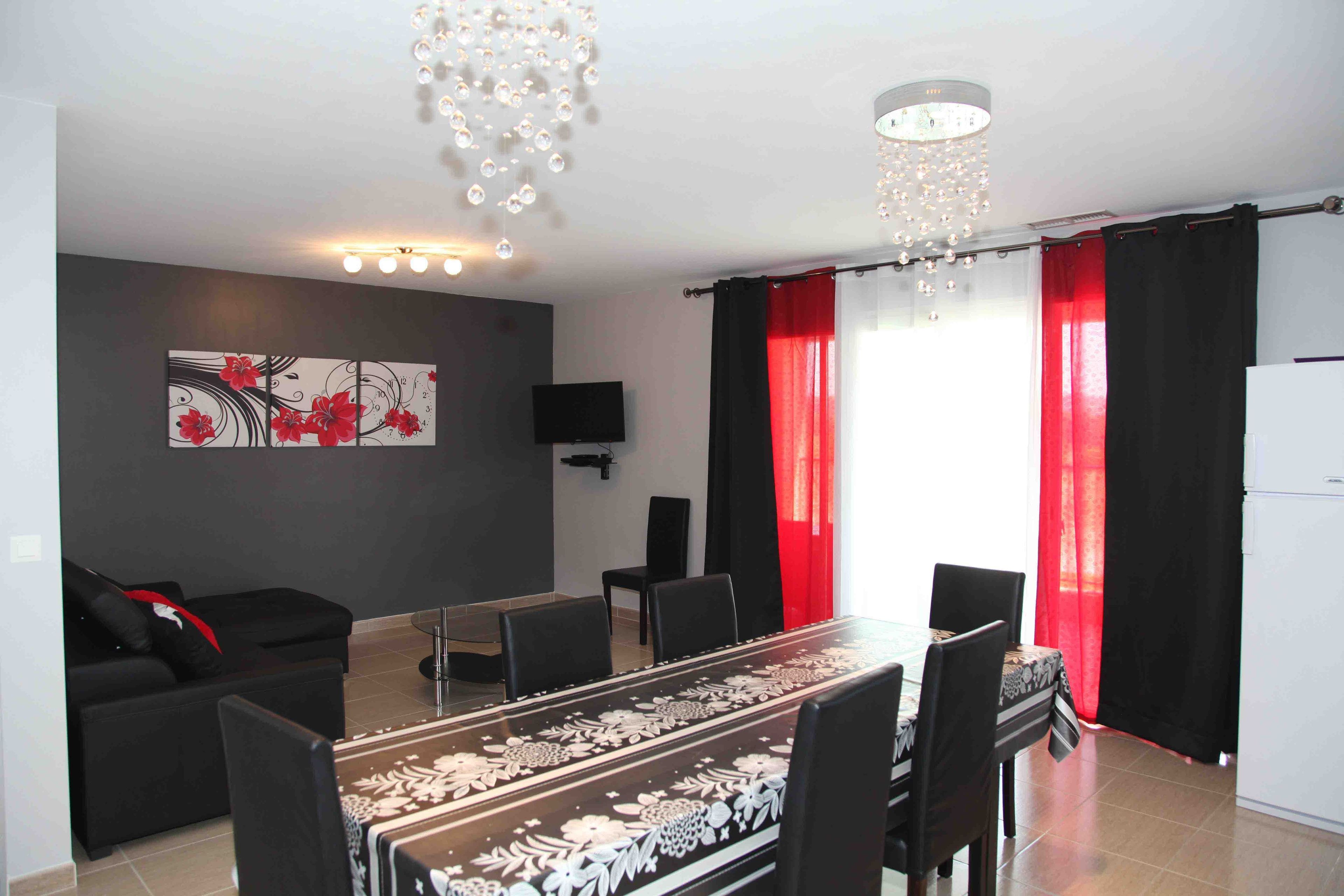 A modern living room with a dining area, a flat-screen TV mounted on the wall, and a chandelier.