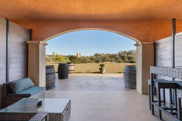 An outdoor patio with a view of a field and distant trees, furnished with wicker furniture and potted plants.