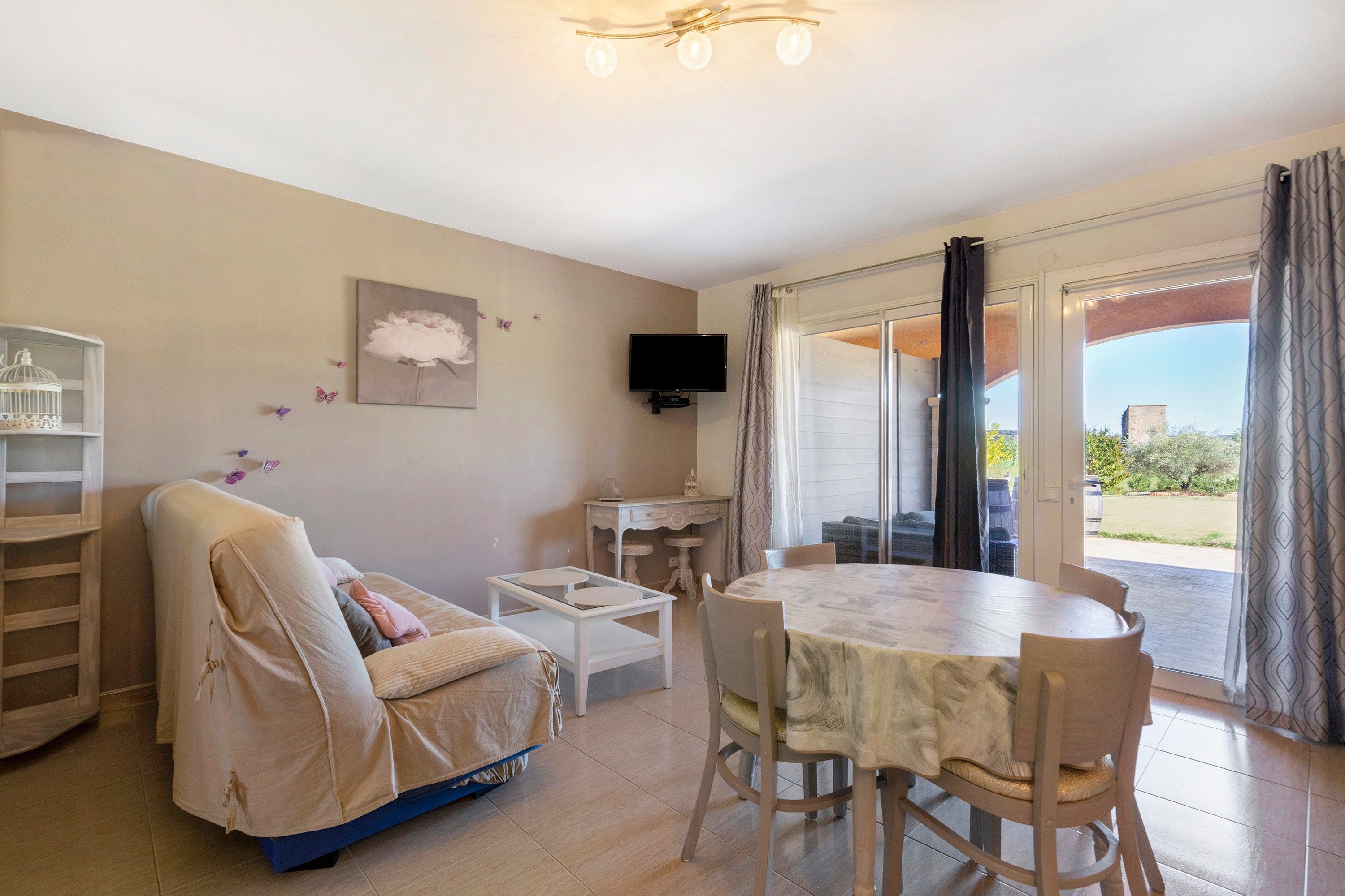 A living room with a sofa, a dining table with chairs, a television, and a balcony with curtains.
