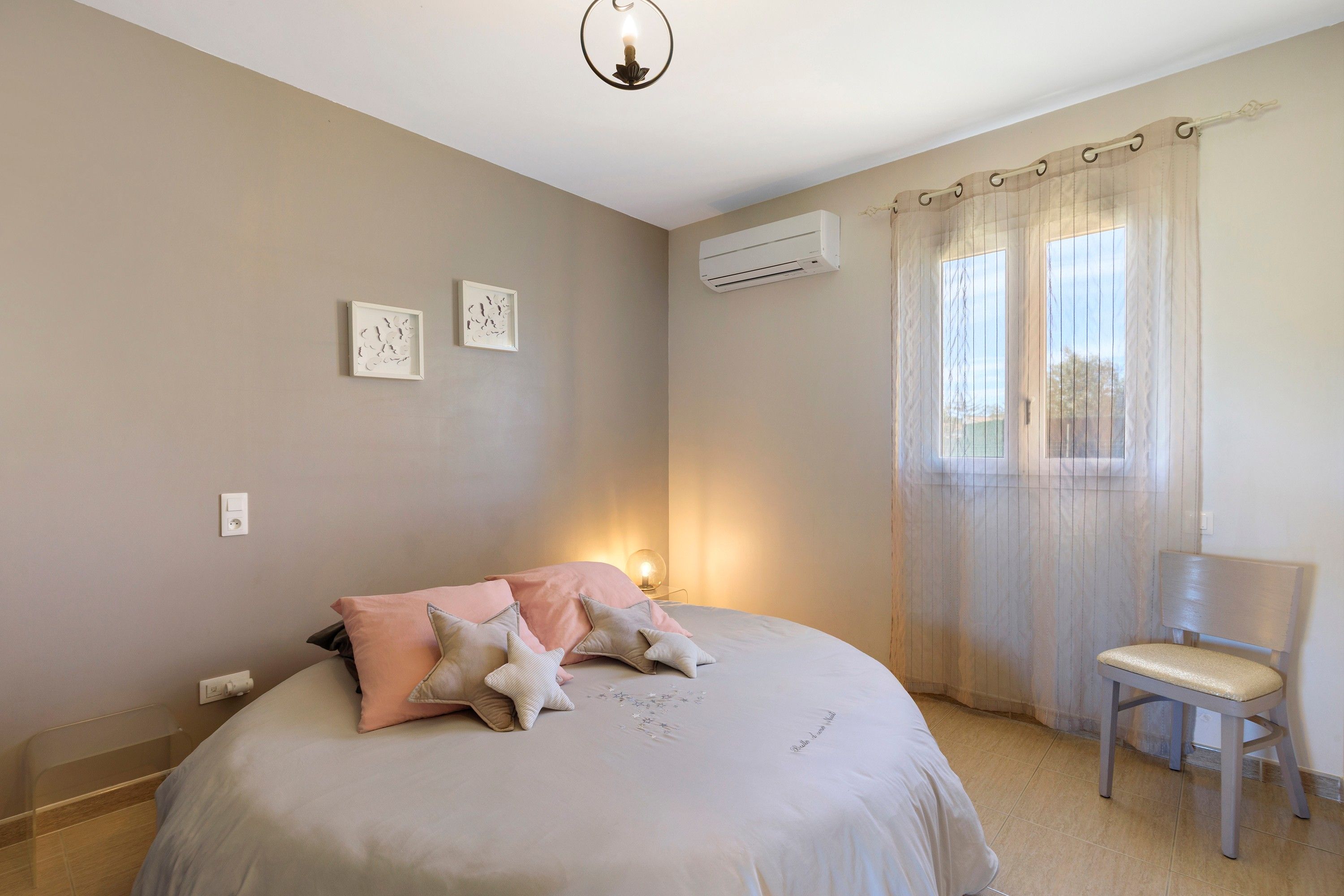 Une chambre à coucher comprenant un lit, une chaise, une fenêtre avec des rideaux, un climatiseur et des œuvres d’art murales.
