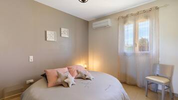 Une chambre à coucher comprenant un lit, une chaise, une fenêtre avec des rideaux, un climatiseur et des œuvres d’art murales.