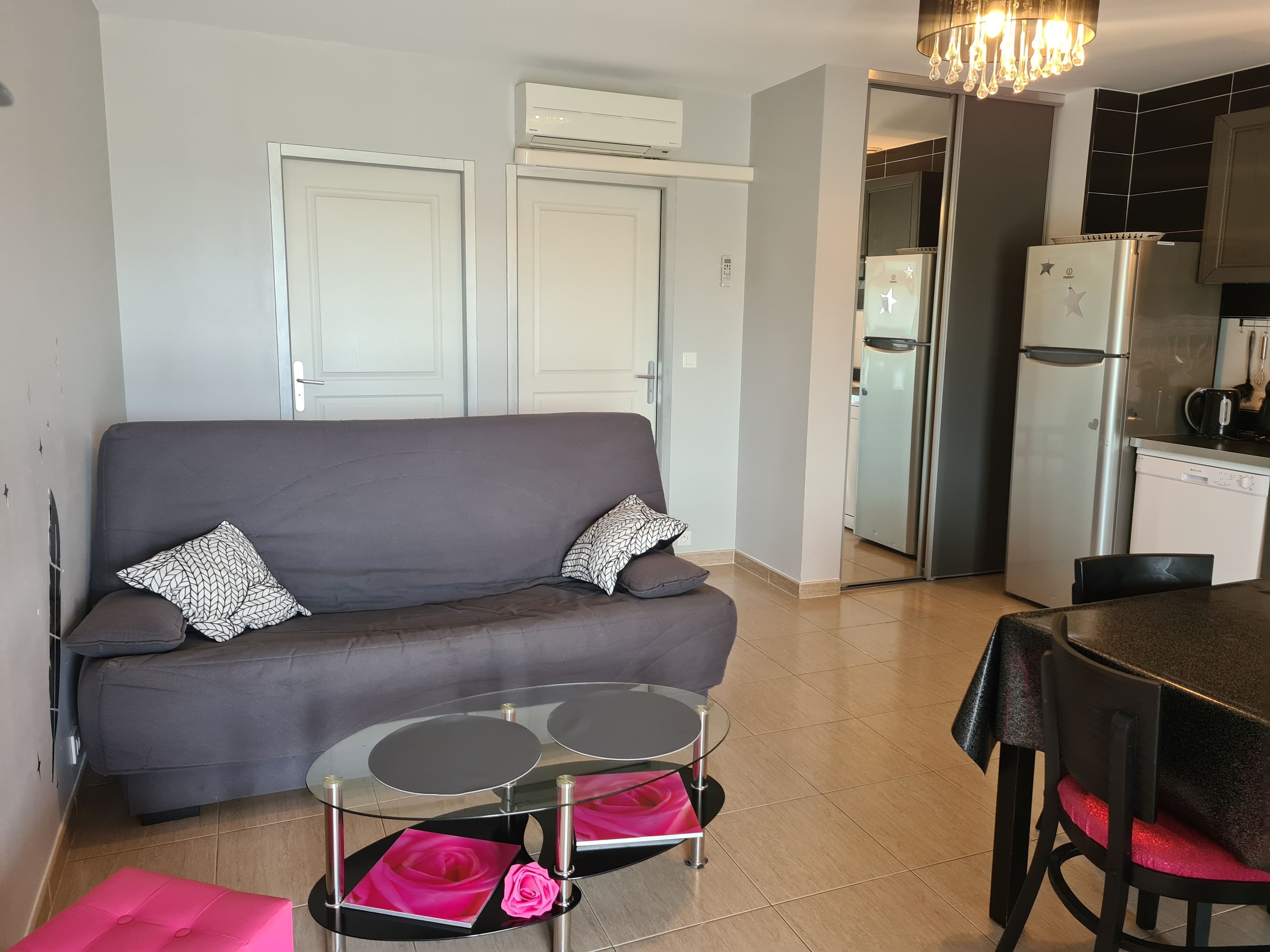 A living room with a grey sofa, a glass coffee table with pink cushions, and a kitchen area with a refrigerator and a dishwasher.