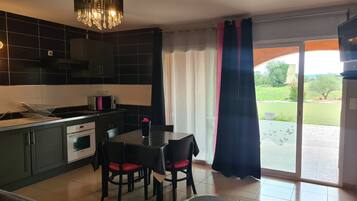 A modern kitchen with dark cabinetry, a dining area with a glass table, and a view of a garden through a sliding glass door.