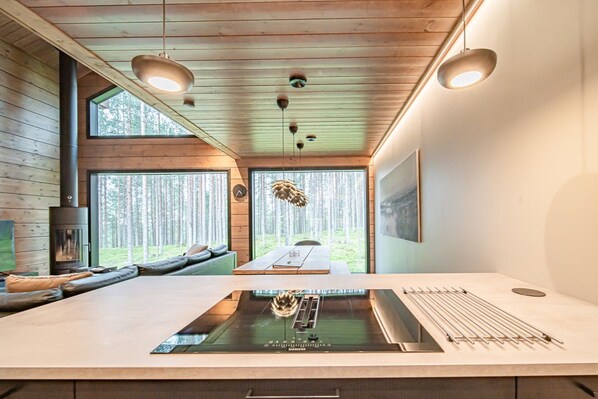 A modern kitchen with a central island, a built-in stove, and a dining area with a view of the forest. - Tieva Chalets Levi by Aavalevi (Levi)