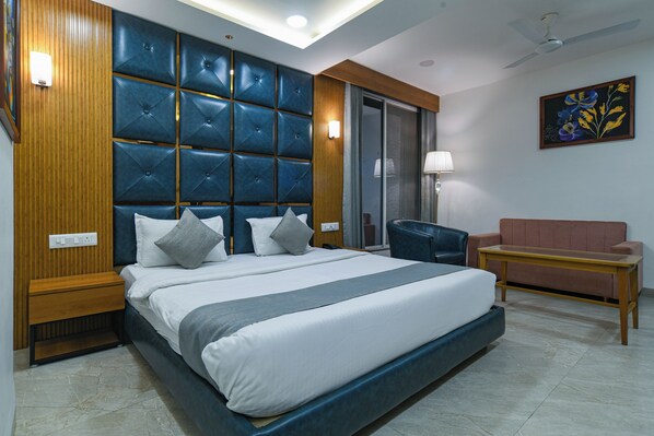 A hotel room with a large bed, a seating area with a sofa and armchair, a painting on the wall, and a ceiling fan.