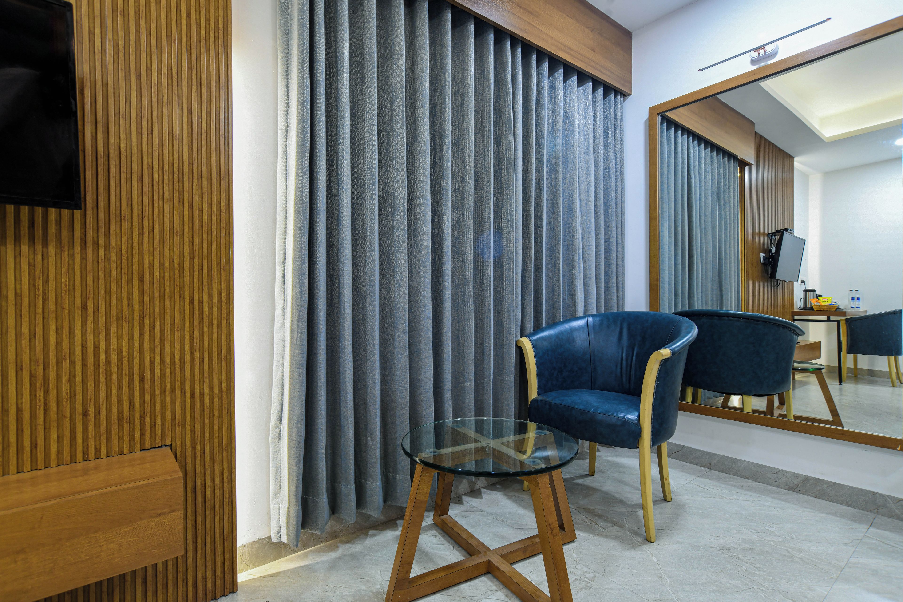 Habitación de hotel con pared de paneles de madera, un sillón azul, una mesita de vidrio y un espejo que refleja la habitación.