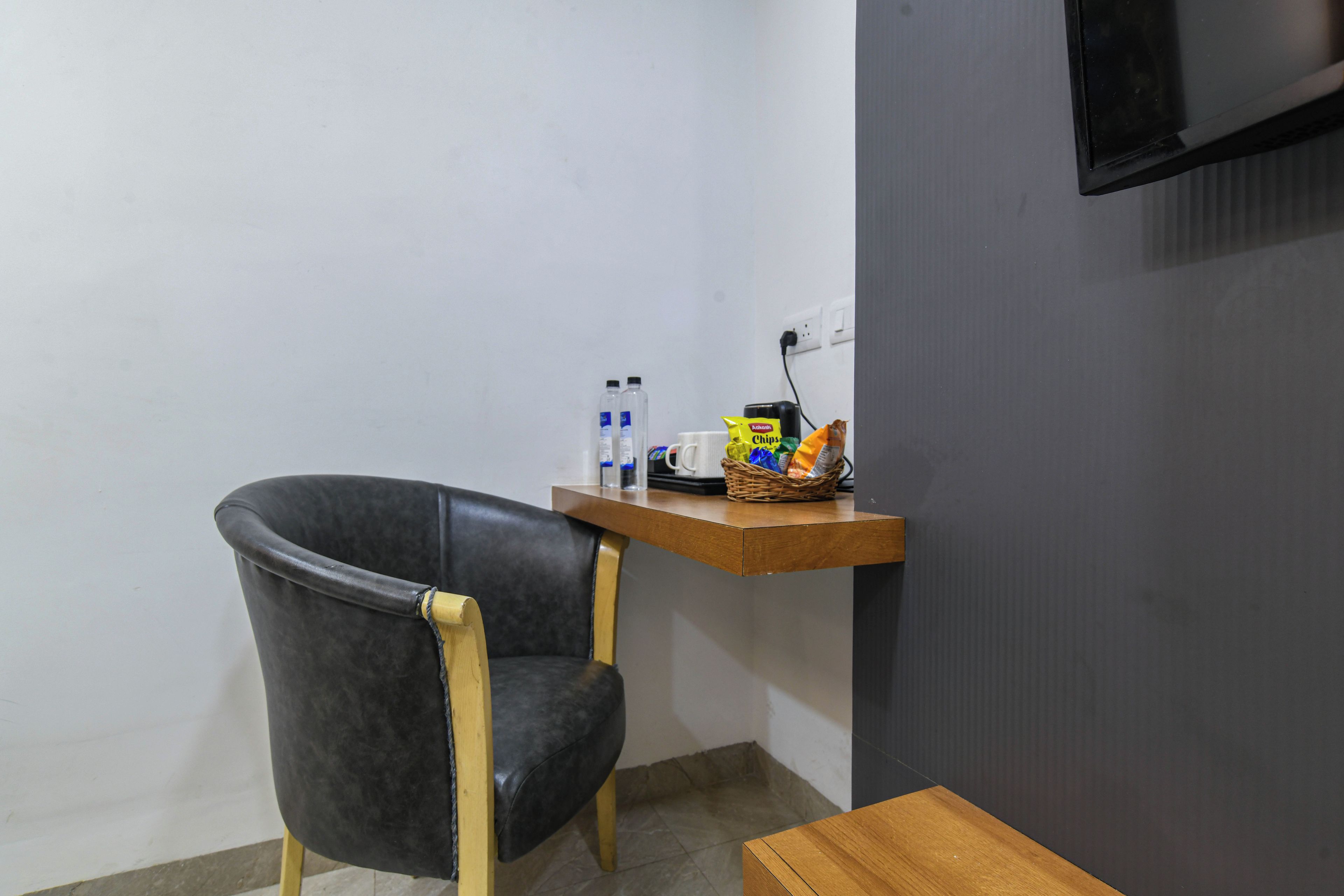 A small room with a black leather chair, a wooden table, a wall-mounted TV, and a shelf with snacks and a kettle.