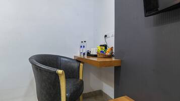 A small room with a black leather chair, a wooden table, a wall-mounted TV, and a shelf with snacks and a kettle.