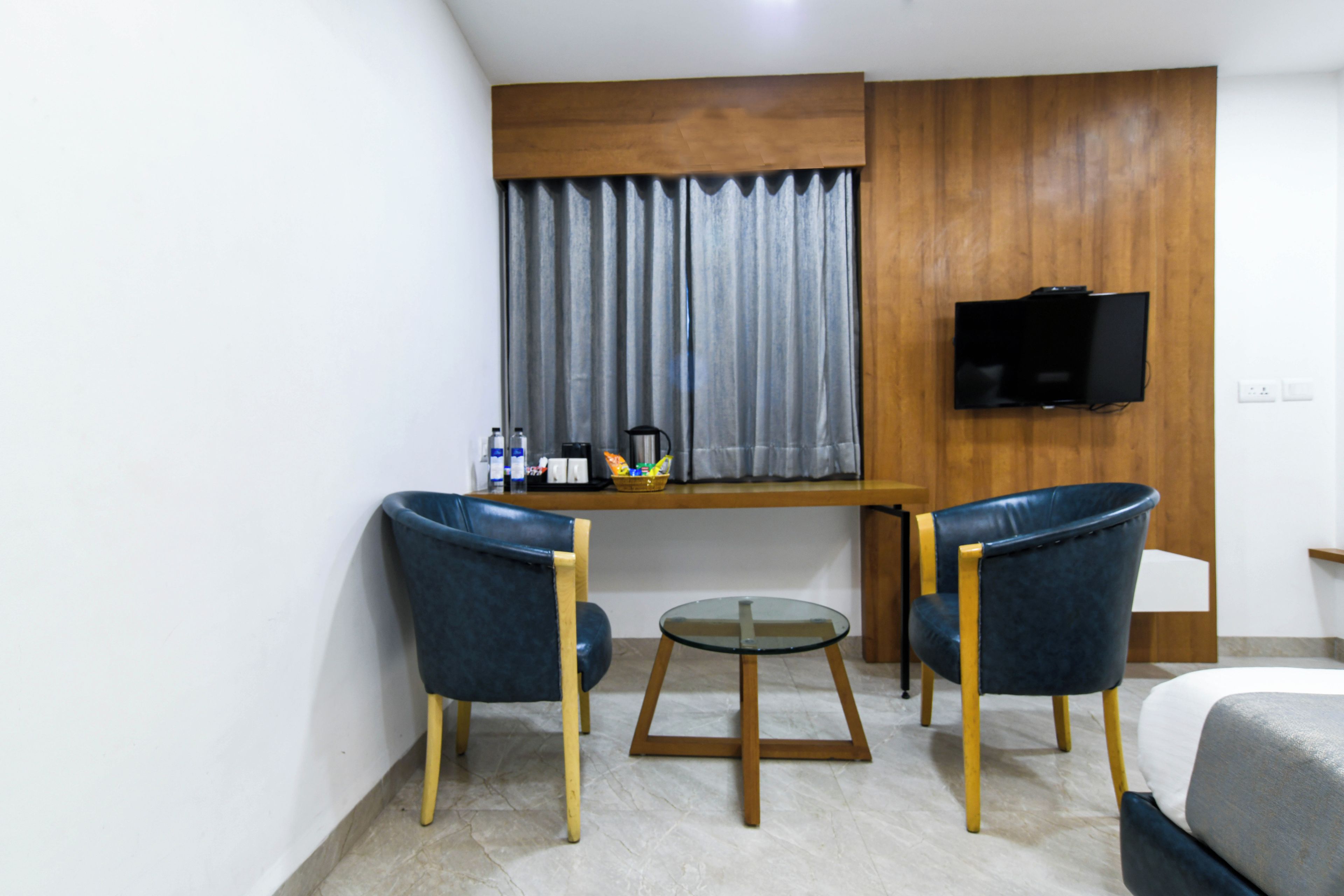 Una habitación de hotel pequeña con cama, dos sillas, una mesita, televisor montado en la pared y ventana con cortinas.