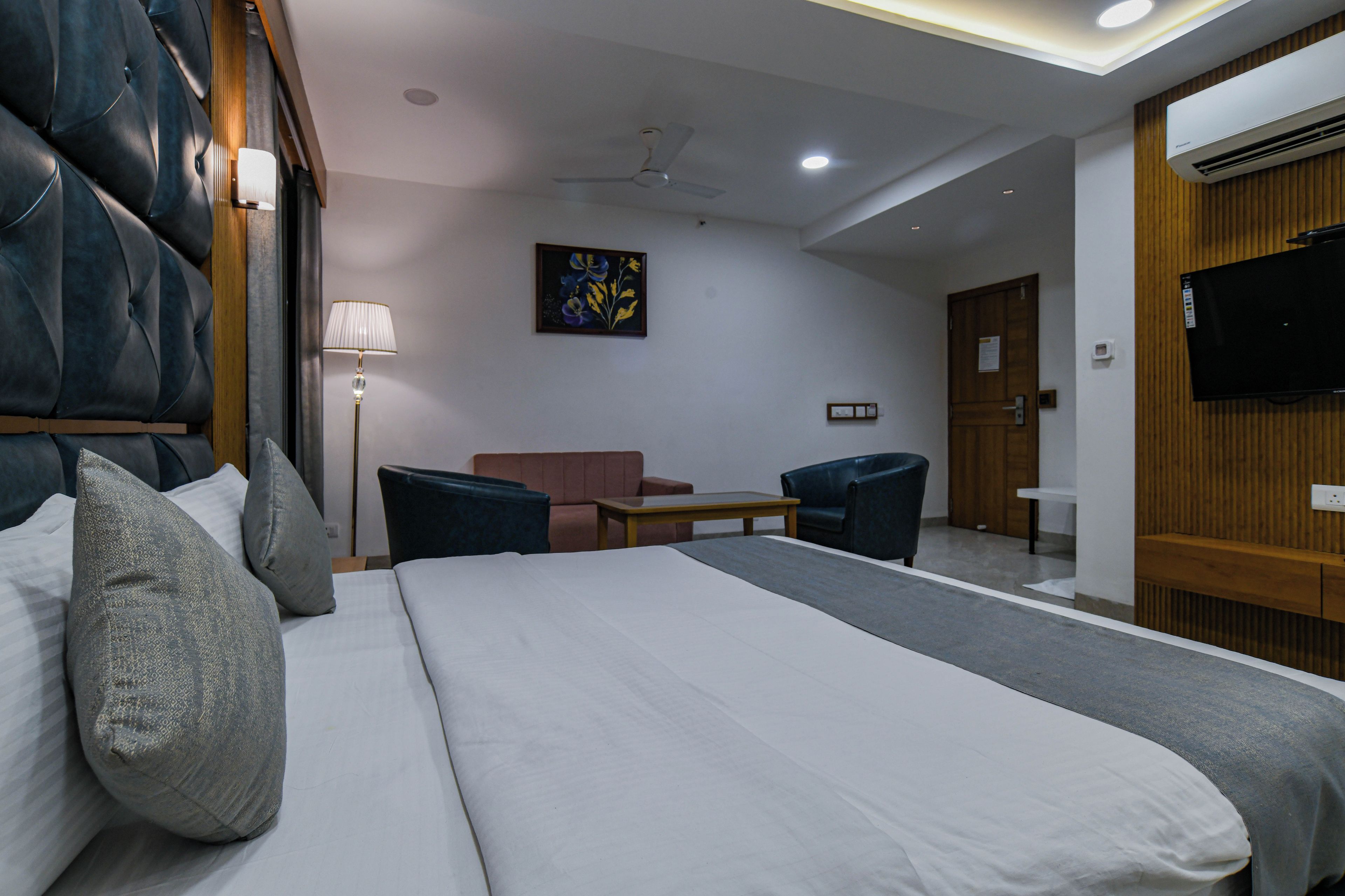 A hotel room with a large bed, a desk, a chair, a sofa, a TV, and an air conditioning unit.