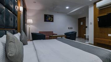 A hotel room with a large bed, a desk, a chair, a sofa, a TV, and an air conditioning unit.