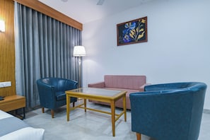 A hotel room with a bed, two blue armchairs, a small table, a pink sofa, a lamp, and a painting on the wall. - Hotel The Grand (Indore)