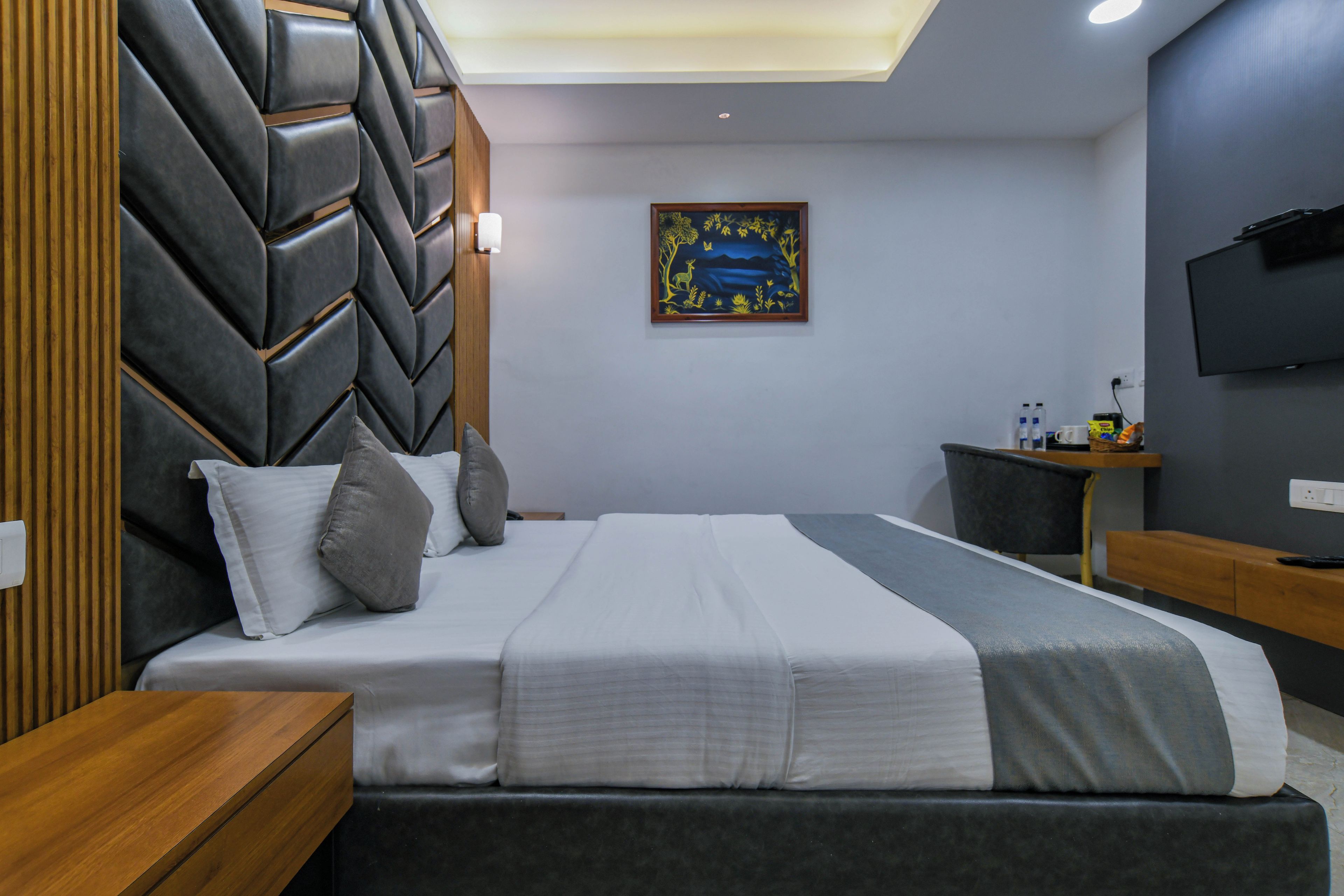 Habitación de hotel moderna con una cama grande, cabecero de madera, mesita de noche pequeña, televisor y un cuadro en la pared.