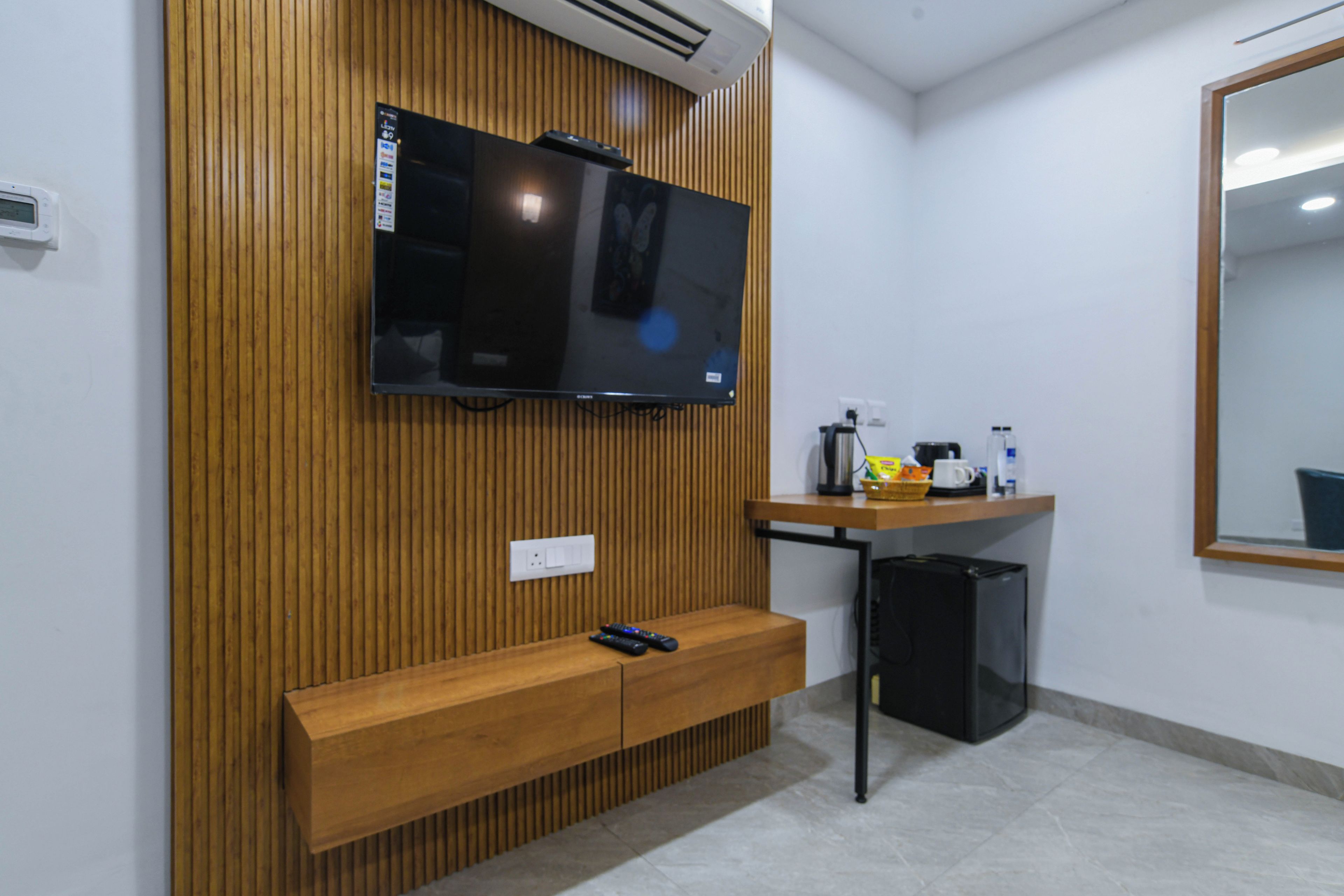 A room with a wooden panel wall, a mounted television, a small wooden bench, a desk with a computer, and a mirror on the wall.