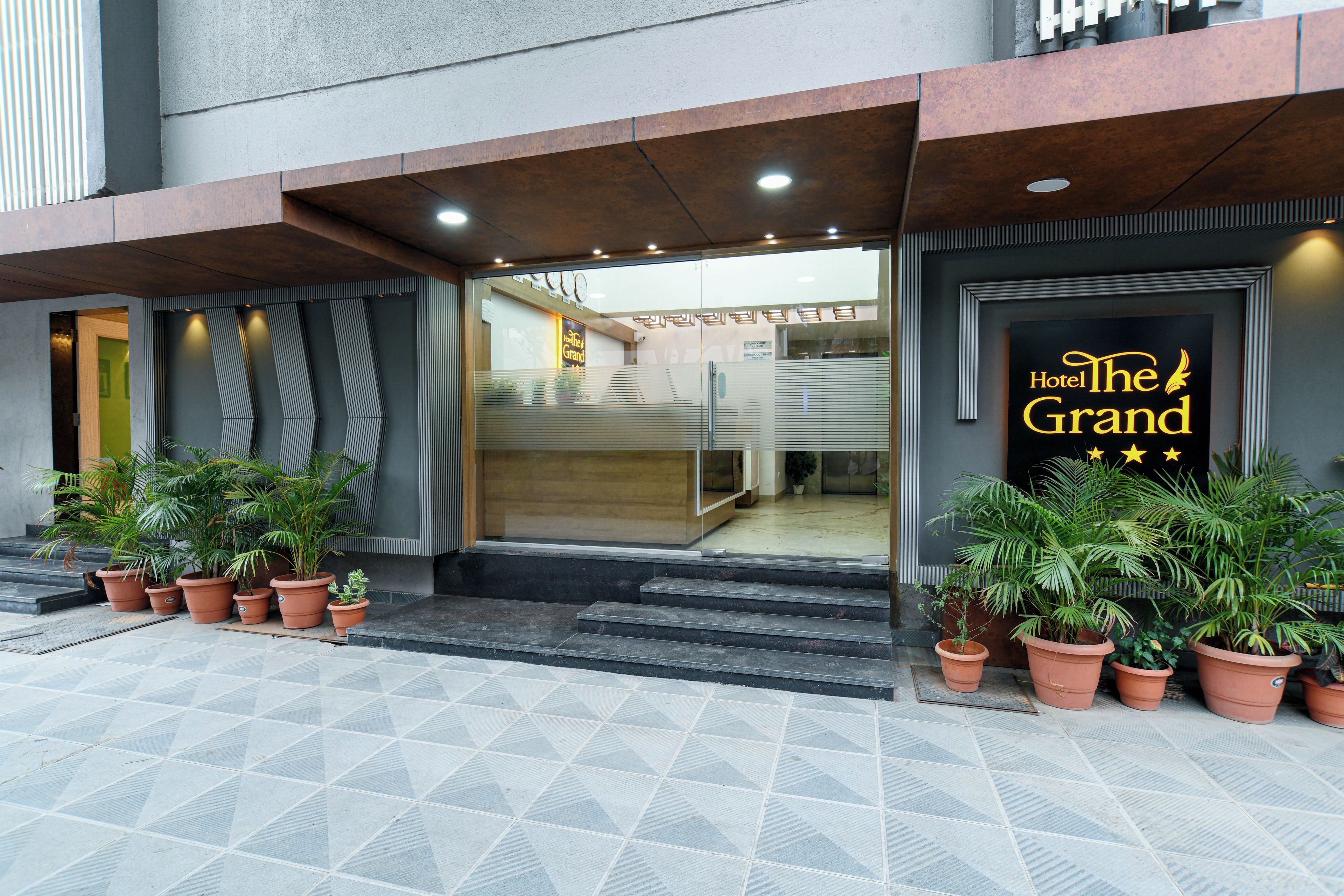 La entrada del Hotel The Grand, con un diseño moderno, presenta un vidrio, plantas en macetas y un letrero con el nombre del hotel.