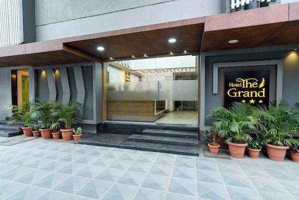 La entrada del Hotel The Grand, con un diseño moderno, presenta un vidrio, plantas en macetas y un letrero con el nombre del hotel.