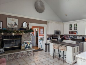 Une cuisine avec des meubles blancs, une cheminée en pierre et un coin repas agrémenté d’un tableau.
