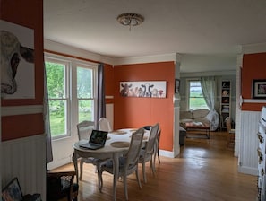 Ein Essbereich mit einem runden Tisch, Stühlen und einem Laptop. An der Wand hängt ein Gemälde und durch das Fenster ist viel Grün zu sehen.