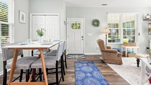 A dining area with a table set for four, a chair, a rug, and a door with a wreath. - Our Little Cottage by the Sea - NEW RENTAL (Pawleys Island)