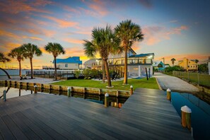 A waterfront property with a wooden deck, palm trees, and a view of the ocean at sunset. - Boat-Friendly Waterfront Luxury Home in Gulf Shores | Sleeps 19 (Gulf Shores)