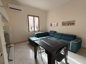 A living room with a sectional sofa, a dining table with chairs, a window with curtains, and two framed pictures on the wall. - Just a short walk from the Lolli station, a quiet and comfortable apartment (palermo)