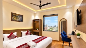 Ein Hotelzimmer mit einem großen Bett, einem Schreibtisch, einem Sessel, einem Fernseher, einem Deckenventilator und einem Fenster mit Blick auf die Stadt.