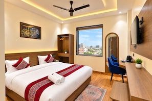 A hotel room with a large bed, a desk, a chair, a TV, a ceiling fan, and a window with a city view. - Hotel Grand Vista (Gorakhpur)