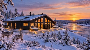 Una cabaña moderna con una amplia terraza, rodeada de árboles cubiertos de nieve y un río.