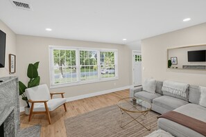 A modern living room with a grey sofa, a white armchair, a coffee table, and a large window with blinds. - Enjoy Quiet Nights With A Fireplace In Bellport 3 Bedroom Home (Bellport)