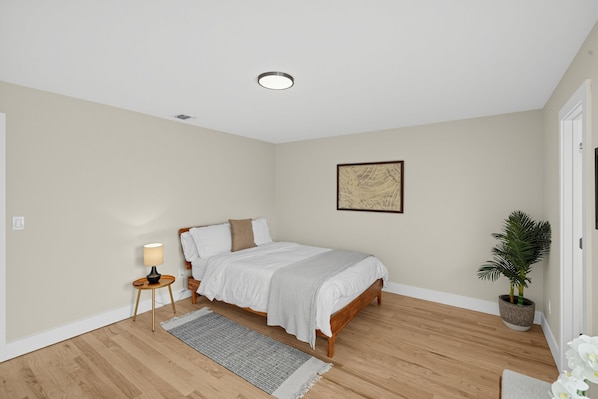 Un dormitorio con cama, una mesita de noche con lámpara, un cuadro en la pared, una planta en maceta y suelo de madera.